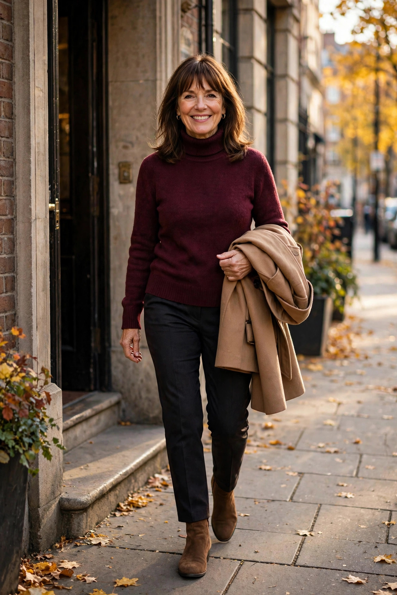Smiling woman with shoulder-length brown hair and bangs in a burgundy turtleneck and dark trousers, carrying a folded camel coat on an autumn sidewalk