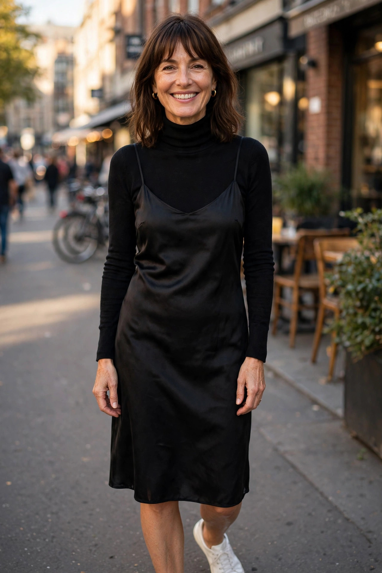 Smiling woman in a black slip dress layered over a black turtleneck with white sneakers, a casual way to style a slip dress for daytime