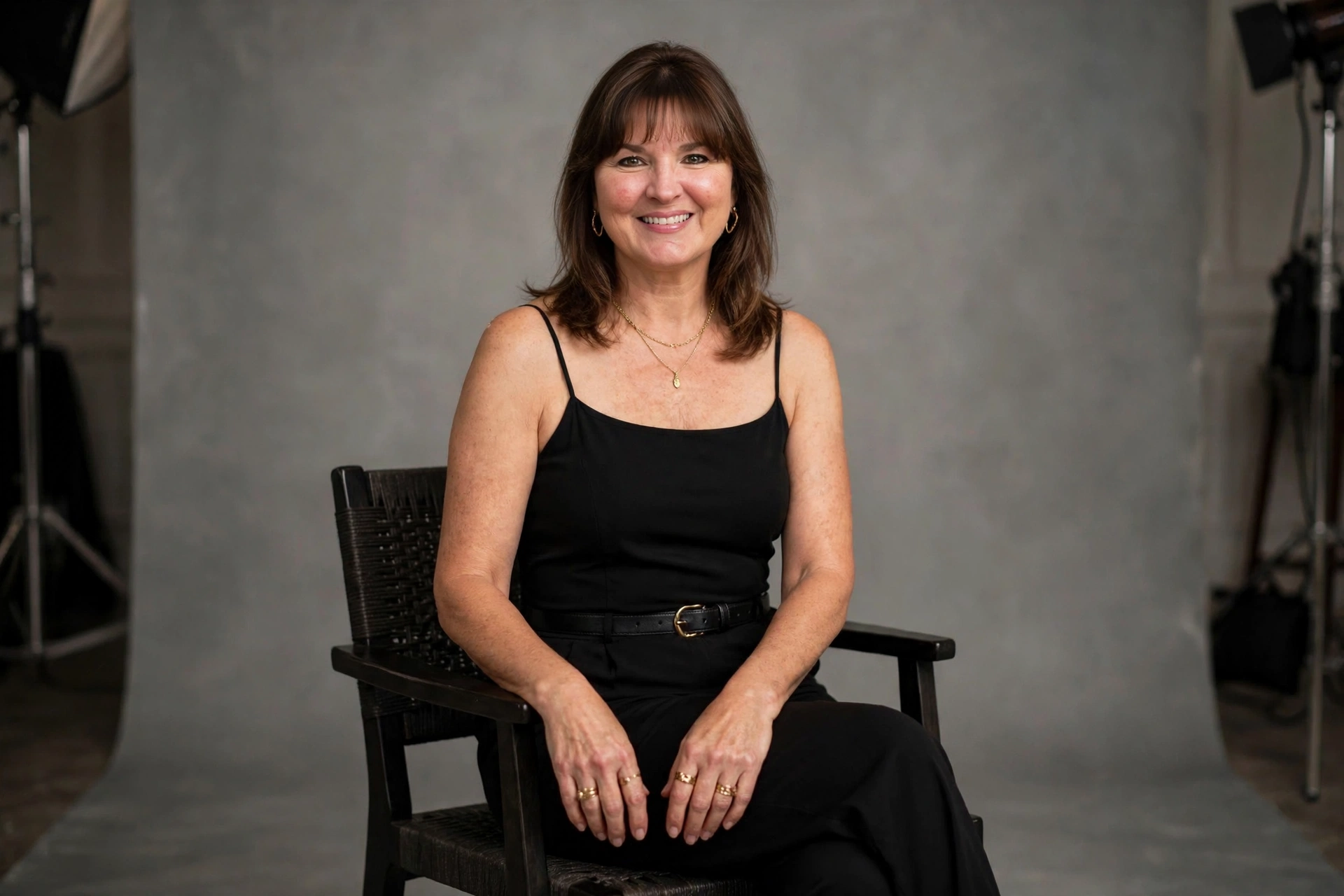 Smiling woman with shoulder-length brown hair and bangs in a black spaghetti-strap jumpsuit with a belt seated on a chair